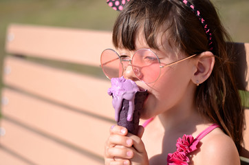 Closeup of pretty little girl eating ice cream outdoors on sunny day. Cute girl in pink swimsuit licking purple ice-cream in waffle cone. Summer, happy childhood concept. Copy space for your text