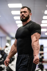 strong bearded sport young man in black sportswear showing his muscle and vein separation on triceps after workout in gym