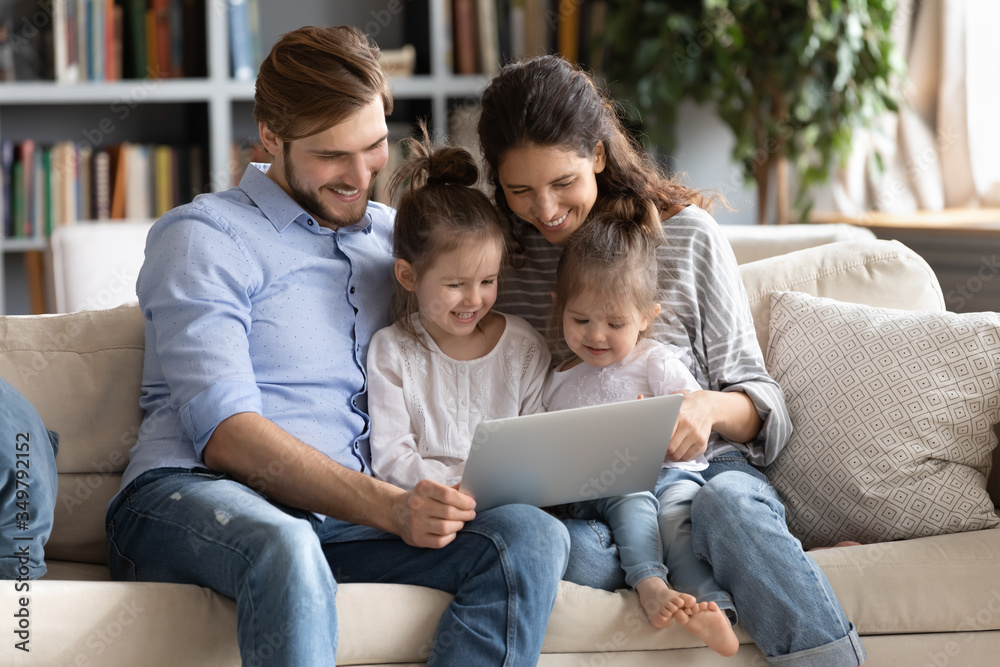 Happy young Caucasian family with small daughters relax on couch at home watching funny video on laptop, smiling parents with little girls children enjoy home weekend using computer gadget together - obrazy, fototapety, plakaty 
