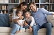 © fizkes - Smiling young Caucasian family with little daughters sit on couch in living room male selfie on smartphone together, happy parents with small children pose for self-portrait picture on cellphone