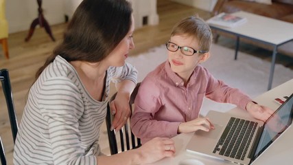Young mother and son are using laptop for education at table in home during quarantine spbas. Beautiful woman and child looking at computer screen with happy smiles while sitting at desk in light room