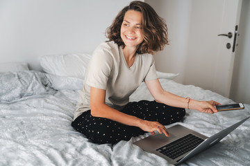 Wall Mural - Pretty young curly woman spends time with a smartphone and a laptop on the bed in the bedroom, remote work education, dates