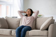 © fizkes - African ethnicity millennial woman in casual clothes puts hands behind head closed eyes resting lean on sofa. Daydream, enjoy fresh conditioned air at modern flat, no stress, reducing fatigue concept