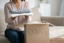 Unrecognizable Mixed-race Woman Cropped Image Close Up View Sit On Couch Near Donation Box Holding Clothes Taking Part At Humanitarian Aid Organization Help To Needy. Old Belongings Charity Concept