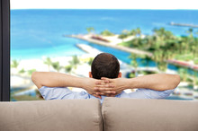 Rear View Of A Single Man Relaxed On A Couch At Home And Looking The Beach Background Outdoors Through The Window In The Living Room At Hotel
