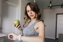 Caucasian Girl With Elegant Hairstyle Looking At Her Watch. Indoor Shot Of Beautiful White Lady With Apple In Hand.