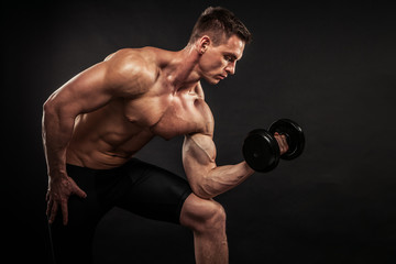 Fitness in gym, sport and healthy lifestyle concept. Handsome athletic man showing his trained body on black background. Bodybuilder male model training biceps muscles with dumbbell.