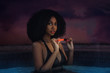 © Phil - A gorgeous young black female with beautiful makeup & long black curly hair posing by herself inside a pool wearing a bikini top and holding an orange lipstick in her hands.