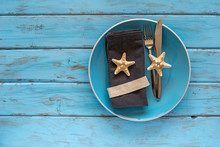Marine Theme Table Setting. Top View Of Blue Plate, Grey Napkins, Cutlery And Star Fishes On Blue Wooden Background With Copy Space.