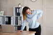 © Admiral - Young office woman with back pain is standing at her desk in front of a laptop, holding her back with her hand