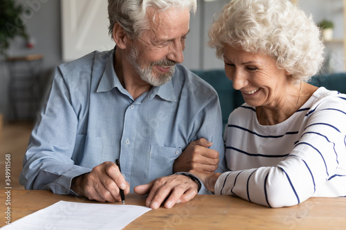 Happy senior older married couple discussing investing saving before signing paper agreement. Smiling middle aged man woman putting signature on marriage contract or making testate at lawyer office.