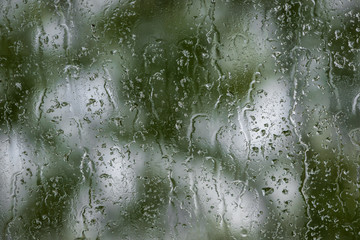 Glass covered with raindrops on a background of blurred greenery. Raindrops flowing down