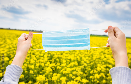 Goodbye tsovid 19! Blue medical disposable face mask in the hands of a child on a background of yellow rapeseed field. Disposable breath filter face mask with earloop