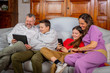 © damianobuffo - Happy family sitting on sofa and using digital tablet and mobile phone at home - Father and son laughing while watching tha tablet and mother and daughter joking in video chat with smartphone