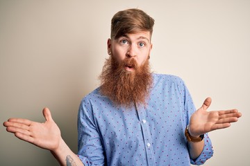 Wall Mural - Handsome Irish redhead business man with beard standing over isolated background clueless and confused expression with arms and hands raised. Doubt concept.