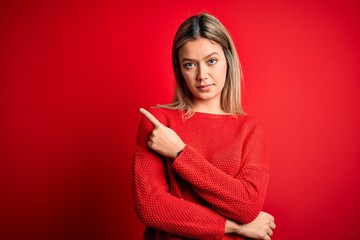 Wall Mural - Young beautiful blonde woman wearing casual sweater over red isolated background Pointing with hand finger to the side showing advertisement, serious and calm face