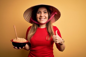 Sticker - Young woman wearing asian hat and eating chinese noodles over yellow isolated background happy with big smile doing ok sign, thumb up with fingers, excellent sign