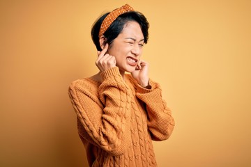 Poster - Young beautiful asian girl wearing casual sweater and diadem standing over yellow background covering ears with fingers with annoyed expression for the noise of loud music. Deaf concept.