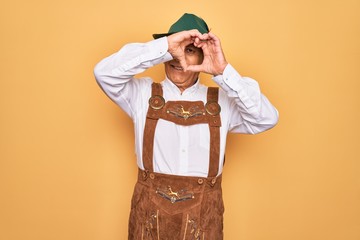 Sticker - Senior grey-haired man wearing german traditional octoberfest suit over yellow background Doing heart shape with hand and fingers smiling looking through sign