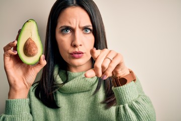 Young woman with blue eyes holding middle healthy avocado over isolated white background pointing with finger to the camera and to you, hand sign, positive and confident gesture from the front