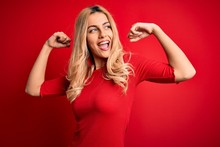 Young Beautiful Blonde Woman Wearing Casual T-shirt Standing Over Isolated Red Background Showing Arms Muscles Smiling Proud. Fitness Concept.