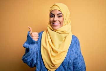 Wall Mural - Young beautiful brunette muslim woman wearing arab hijab over isolated yellow background doing happy thumbs up gesture with hand. Approving expression looking at the camera showing success.