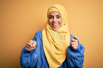 Wall Mural - Young beautiful brunette muslim woman wearing arab hijab over isolated yellow background pointing fingers to camera with happy and funny face. Good energy and vibes.