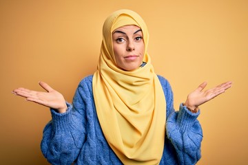 Wall Mural - Young beautiful brunette muslim woman wearing arab hijab over isolated yellow background clueless and confused with open arms, no idea concept.