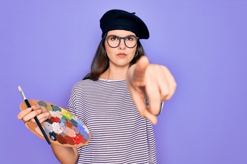 Sticker - Young beautiful artist girl wearing fashion beret painting using paintbrush and palette pointing with finger to the camera and to you, hand sign, positive and confident gesture from the front