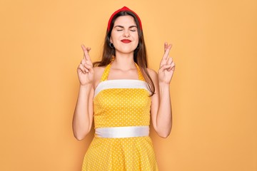 Poster - Young beautiful pin up woman wearing 50s fashion vintage dress over yellow background gesturing finger crossed smiling with hope and eyes closed. Luck and superstitious concept.