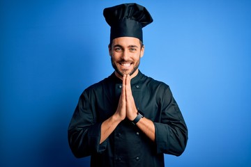 Canvas Print - Young handsome chef man with beard wearing cooker uniform and hat over blue background praying with hands together asking for forgiveness smiling confident.