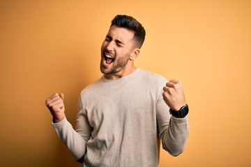 Wall Mural - Young handsome man wearing casual sweater standing over isolated yellow background very happy and excited doing winner gesture with arms raised, smiling and screaming for success. Celebration concept.