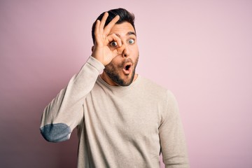 Sticker - Young handsome man wearing casual sweater standing over isolated pink background doing ok gesture shocked with surprised face, eye looking through fingers. Unbelieving expression.