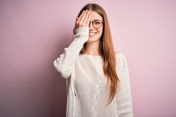 Young beautiful redhead woman wearing casual sweater and glasses over pink background covering one eye with hand, confident smile on face and surprise emotion.