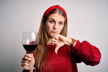 Wall Mural - Young beautiful redhead woman drinking glass of red wine over isolated white background with angry face, negative sign showing dislike with thumbs down, rejection concept