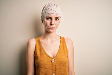 Young beautiful redhead woman injured for accident wearing bandage on head with serious expression on face. Simple and natural looking at the camera.