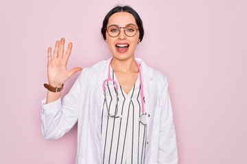 Wall Mural - Beautiful doctor woman with blue eyes wearing coat and stethoscope over pink background showing and pointing up with fingers number five while smiling confident and happy.