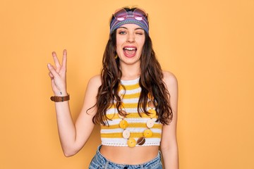 Wall Mural - Young beautiful hippie woman with blue eyes wearing accesories and sunnglasses smiling with happy face winking at the camera doing victory sign with fingers. Number two.