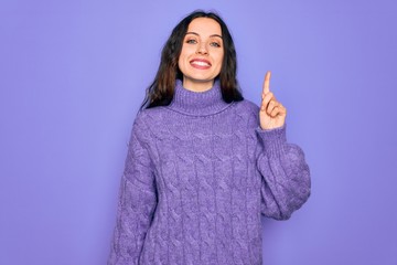 Wall Mural - Young beautiful woman wearing casual turtleneck sweater standing over purple background showing and pointing up with finger number one while smiling confident and happy.