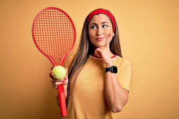 Wall Mural - Beautiful brunette sportswoman playing tennis using racket and ball over yellow background serious face thinking about question, very confused idea