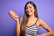 © Krakenimages.com - Young woman eating hotdog with ketchup and mustard over purple background with a happy face standing and smiling with a confident smile showing teeth