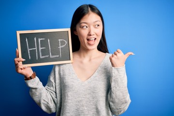 Young asian woman holding blackboard with help text as support message over blue background pointing and showing with thumb up to the side with happy face smiling