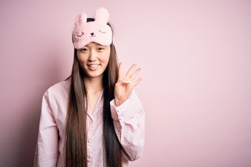 Sticker - Young asian woman wearing pajama and sleep mask over pink isolated background showing and pointing up with fingers number four while smiling confident and happy.