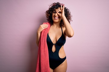 Poster - Young beautiful curly arab woman on vacation wearing swimsuit holding beach towel with happy face smiling doing ok sign with hand on eye looking through fingers