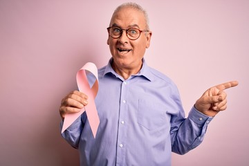 Wall Mural - Middle age hoary man holding pink cancer ribbon symbol over isolated background very happy pointing with hand and finger to the side