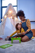 © Svitlana - A babysitter you can trust. African american woman baby sitter entertaining caucasian cute little girl. Kid reading a book with her nanny sitting in wigwam, tent