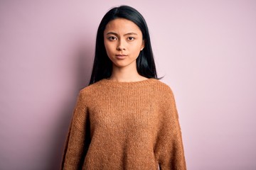 Young beautiful chinese woman wearing casual sweater over isolated pink background with serious expression on face. Simple and natural looking at the camera.