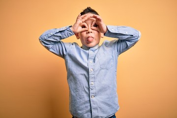 Poster - Young little boy kid wearing elegant shirt standing over yellow isolated background doing ok gesture like binoculars sticking tongue out, eyes looking through fingers. Crazy expression.