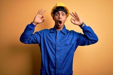 Wall Mural - Young handsome african american worker man wearing blue uniform and security helmet looking surprised and shocked doing ok approval symbol with fingers. Crazy expression