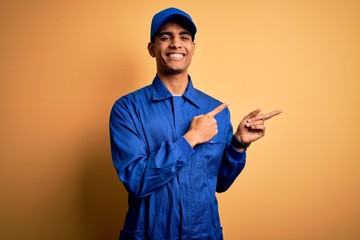 Wall Mural - Young african american mechanic man wearing blue uniform and cap over yellow background smiling and looking at the camera pointing with two hands and fingers to the side.
